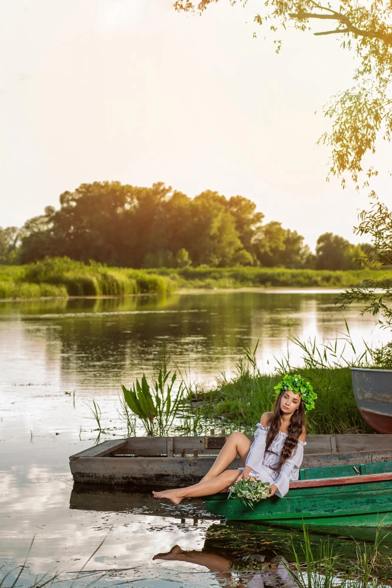 Junge Frau mit Blumenkranz im Haar, entspannt bei Sonnenuntergang in einem Boot auf einem Fluss. Schöner Körper und schönes Gesicht. Fantasy-Kunstfotografie. Konzept weiblicher Schönheit, Erholung im Dorf.